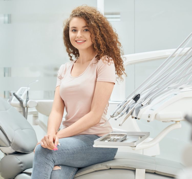 Beautiful young girl with curly hair wearing pink shirt and blue jeans sitting in dental chair after doctor's appointment. Female patient visiting professional dentist at modern clinic.